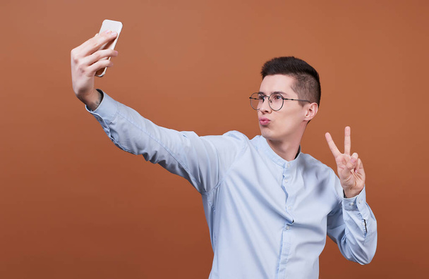 A cute, cheerful, joyful man, wearing glasses and a blue shirt, holds a mobile phone in his hands and takes a selfie. Photo on a brown background. - Fotó, kép