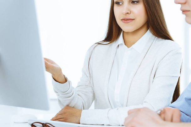 Молодая женщина сидит за столом с компьютером в белом офисе. Похоже, студентка или деловая леди общаются со случайным одетым мужчиной - Фото, изображение