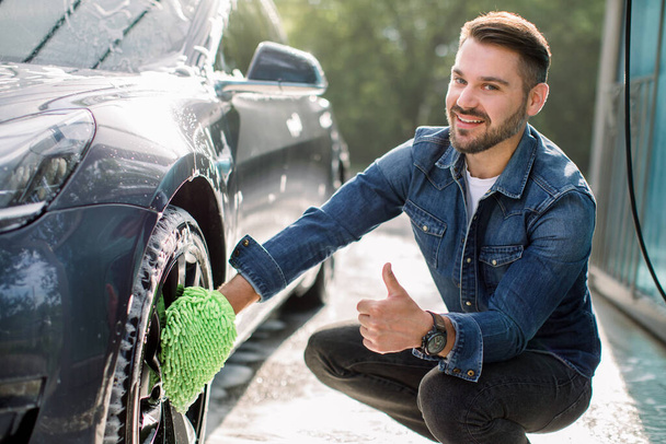 Smiling handsome young man showing thumb up and looking at camera, while using green wash mitt for cleaning rims of his modern car outdoors. Luxury blue electric car in soap foam outdoors at car wash - Foto, imagen