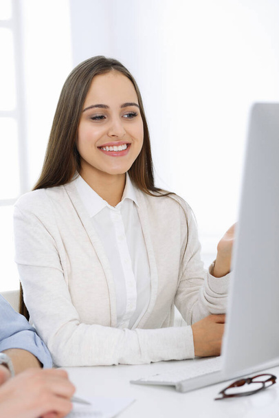 Young woman sitting at the desk with computer in white colored office. Looks like student girl or business lady communicating with casual dressed man - Φωτογραφία, εικόνα