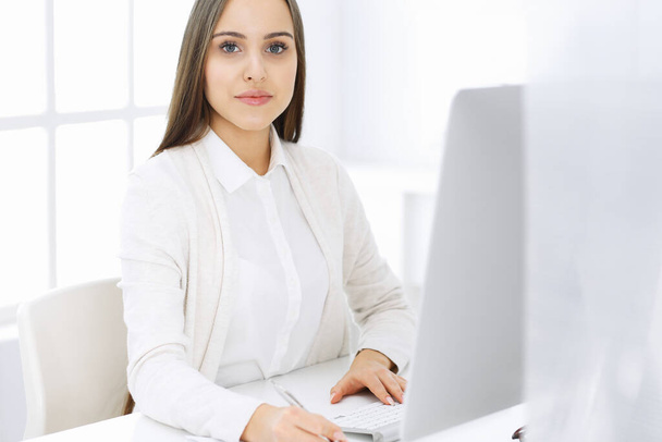 Young cheerful woman sitting at the desk with computer and looking at camera in white colored office. Looks like student girl or business lady. Headshot - Foto, imagen
