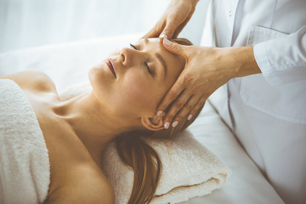 Beautiful happy woman enjoying facial massage with closed eyes in spa salon. Relaxing treatment in medicine and Beauty concept - Foto, afbeelding