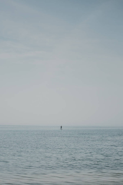 Minimaalinen merimaisema, jossa on tyyntä vettä ja joku horisontin lähellä nauttimassa aamusta seisaaltaan melalla. Rentouttavaa ja hauskaa ulkona kesäaktiviteetteja matkailijoille. Meriliikenne aktiivisille ihmisille. - Valokuva, kuva