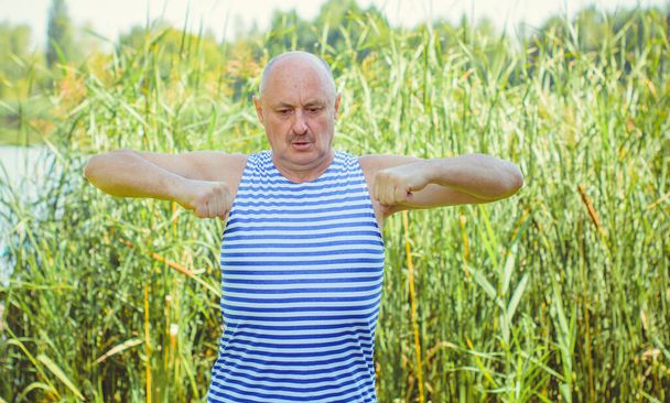 Senior man do exercises on a nature. Physiotherapy concept. Elderly man practicing sports on the street. Full length of mature man stretching muscles during exercise against reeds. - Photo, Image