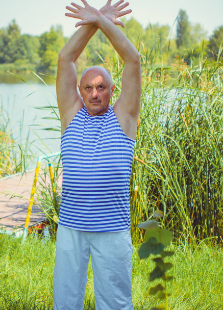 Senior man do exercises on a nature. Physiotherapy concept. Elderly man practicing sports on the street. Full length of mature man stretching muscles during exercise against reeds. - Foto, imagen