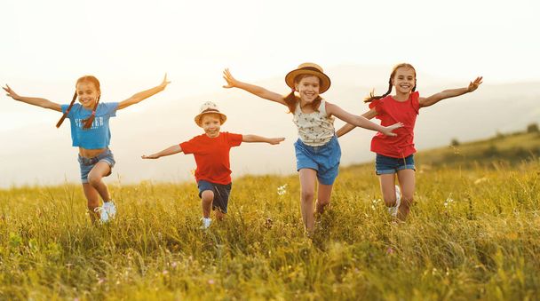 Веселые маленькие дети в повседневной одежде веселятся и бегут вместе в зеленом поле на закате, наслаждаясь летними каникулами в сельской местности - Фото, изображение