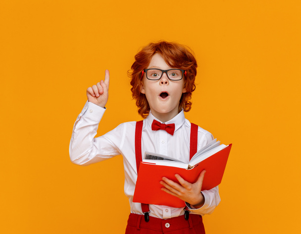 Chytrý zázvorový chlapec v brýlích ukazuje nahoru při čtení učebnice během studia na žlutém pozadí - Fotografie, Obrázek
