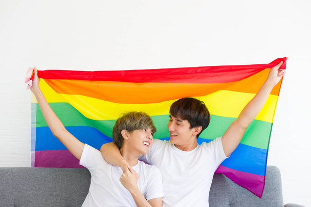 Szczęśliwi azjatyccy homoseksualiści lub pary gejowskie ściskają się razem z tęczową flagą siedzącą na kanapie. Koncepcja dumy LGBTQ. - Zdjęcie, obraz