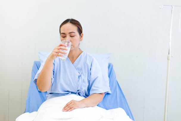 Die Asiatin im Kleid der Patientin hatte Durst. Der Patient trinkt Wasser während er auf dem Bett sitzt. - Foto, Bild