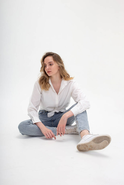 caucasian woman posing in shirt and blue jeans, sitting on white studio floor - Foto, imagen