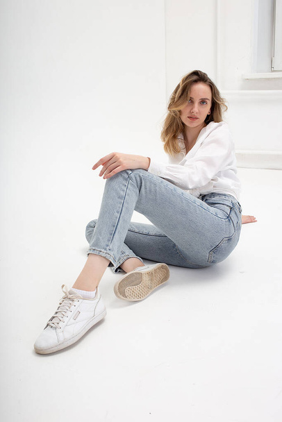 caucasian woman posing in shirt and blue jeans, sitting on white studio floor - Foto, afbeelding