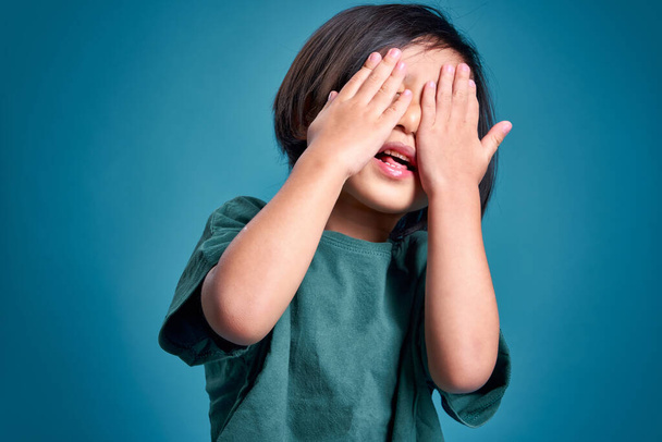 Szczęśliwy azjatycki dzieciak blokujący twarz obiema rękami, puste miejsce w studiu, odizolowany na kolorowym niebieskim tle. Koncepcja edukacji szkolnej. - Zdjęcie, obraz