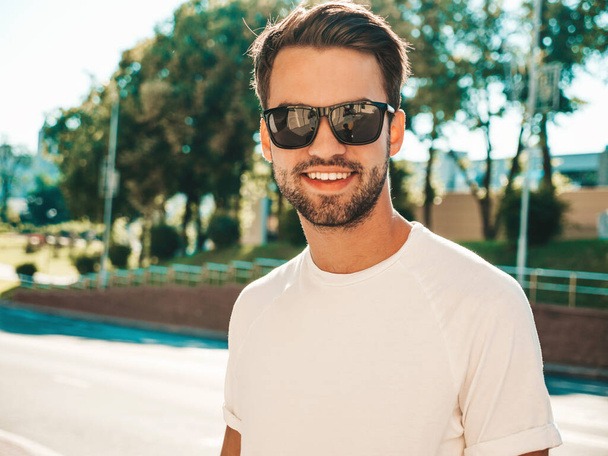 Portrait of handsome smiling stylish hipster lambersexual model.Man dressed in white T-shirt. Fashion male posing on the street background in sunglasses - Φωτογραφία, εικόνα