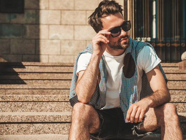 Portrait of handsome confident stylish hipster lambersexual model.Man dressed in white T-shirt. Fashion male sitting at the stairs on the street background in sunglasses - Fotografie, Obrázek