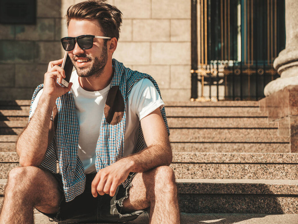Handsome smiling stylish hipster lambersexual model.Man dressed in white T-shirt.Fashion male posing in the street. Holding, speaking  and chatting in his cellphone. Discussing his good day - 写真・画像
