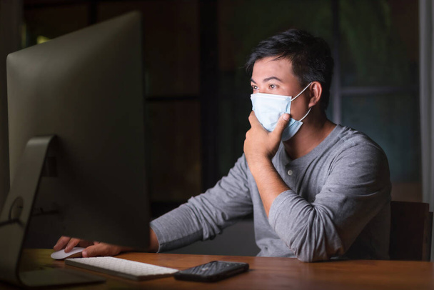 Asiatischer Mann mit Schutzmaske, der nachts von zu Hause aus an der Coronavirus-Krankheit (COVID-19) arbeitet. Arbeit von zu Hause aus und gesundes Konzept - Foto, Bild