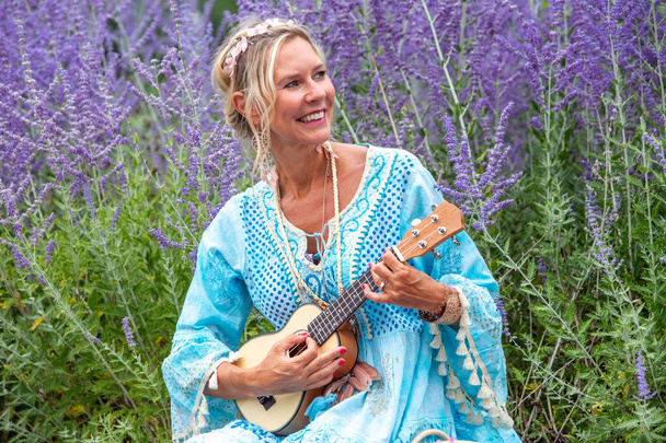 blond woman in her 40s sitting at field of flowers and playing the guitar - Fotó, kép