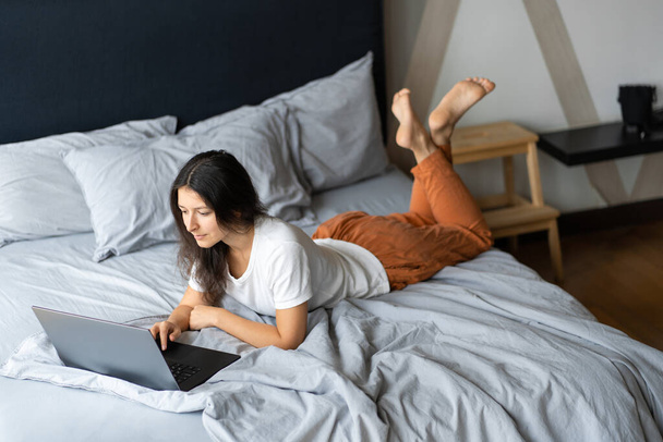 Beautiful young brunette girl with a laptop lying on the bed. Stylish modern interior. A cozy workplace. Shopping on the Internet - 写真・画像