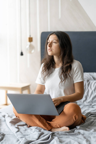 Beautiful young brunette girl with a laptop sitting on the bed. Stylish modern interior. A cozy workplace. Shopping on the Internet. - Foto, Imagem