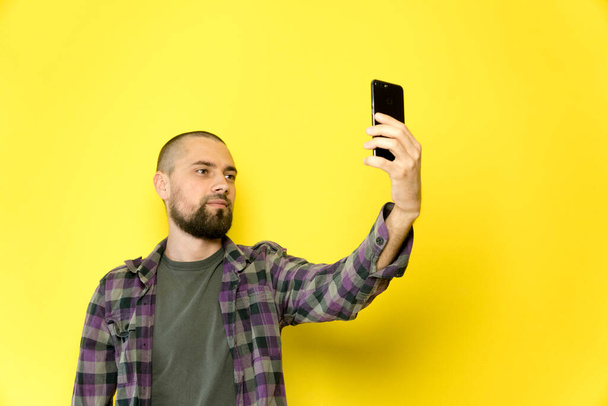Ein junger Mann mit Bart und Glatze hält auf gelbem Grund ein Handy in der Hand. Blogger macht ein Selfie - Foto, Bild