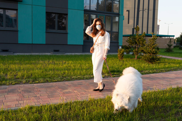 A girl in a dress and a protective mask on her face walks with a white furry dog in a residential area - Фото, изображение