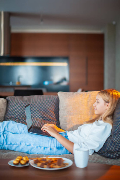 Bella ragazza adolescente sdraiata sul divano con computer portatile a schermo bianco a guardare serie TV con dolci e tè. Ambiente domestico accogliente - Foto, immagini