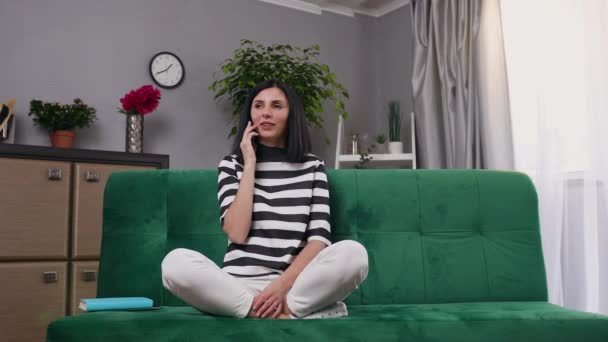 Beautiful smiling lucky young woman with dark hair in stylish clothes sitting on couch in relaxing pose and talking on phone,4k - Felvétel, videó