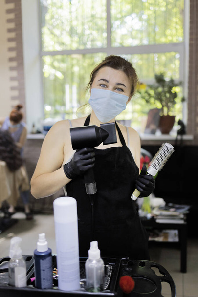 Hairdressing services during the coronavirus. Portrait of girl hairdresser in medical mask and gloves holds comb and hairdryer - Photo, Image