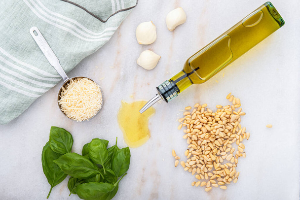 Fotografie čerstvých složek bazalkového pesta včetně panenského olivového oleje, piniových ořechů, česneku, bazalky a parmezánu-Reggiano sýra s kuchyňským ručníkem na pultu - Fotografie, Obrázek