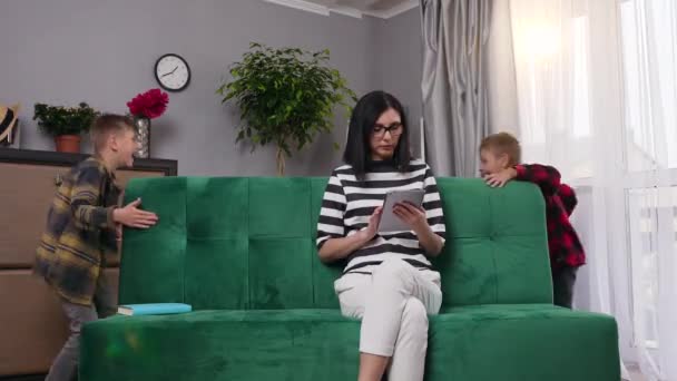 Appealing concentrated tired young woman in glasses working with tablet pc while two noisy loud sons interfering her and running around sofa on which she sits,4k - Imágenes, Vídeo