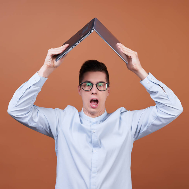 An image of a young funny dark-haired guy in a blue shirt and round glasses for vision, holding a laptop over his head. The young man makes funny faces, a cheerful student. - Fotoğraf, Görsel