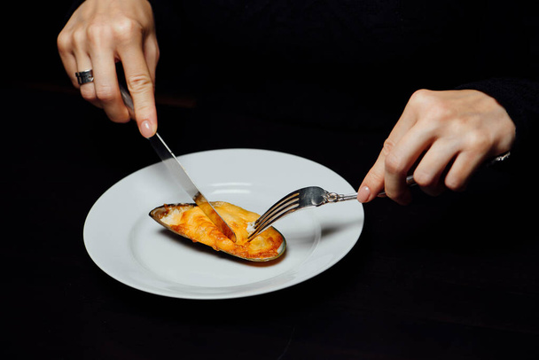 baked mussel in a shell with cheese on a white plate with a knife and fork on a black background - Fotografie, Obrázek