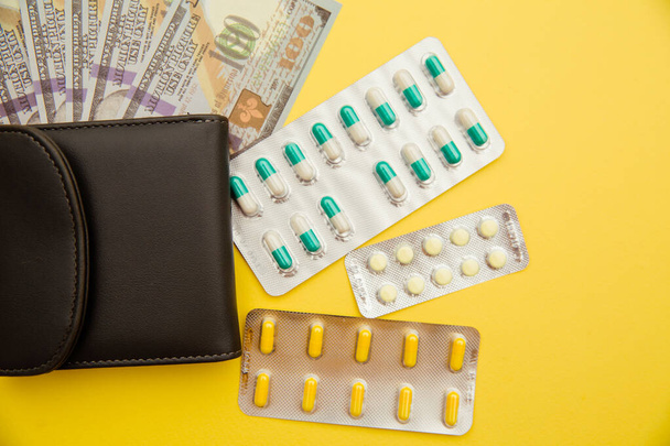 Grey wallet with money and tablets of pills isolated on yellow background - Foto, Imagen