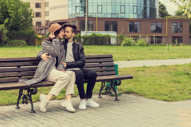 Love story of young couple sitting on the bench and kissing, hugging. Lovely man and woman in the city park wear trendy stylish clothes. Image with copy space. - Фото, изображение