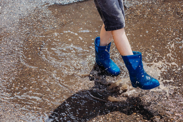 Dzieci latem po deszczu. Zbliżenie stóp chłopca w niebieskich gumowych butach skaczących w kałużach na spacerze. Dzieci bawią się na świeżym powietrzu w deszczową pogodę. Buty wodoodporne dla chłopców. - Zdjęcie, obraz