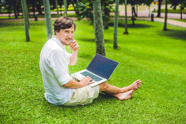 Man businessman or student in casual dress using laptop in a tropical park on the background of skyscrapers. Dressing in a white shirt, beige shorts. Mobile Office concept - Φωτογραφία, εικόνα