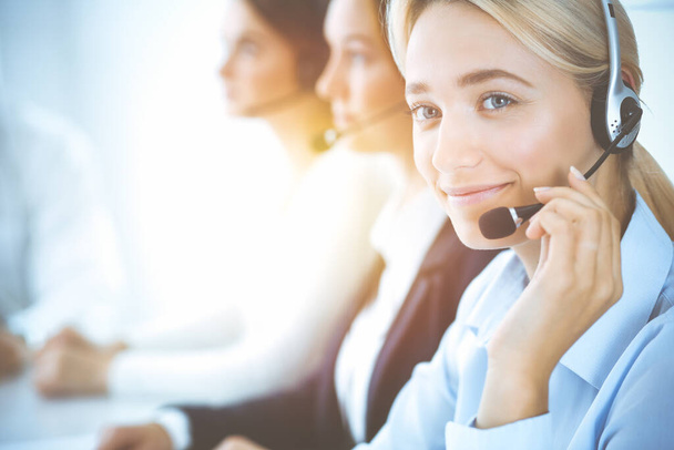 Cheerful smiling business woman with headphones consulting clients. Group of diverse phone operators at work in sunny office.Call center and business people concept - Φωτογραφία, εικόνα