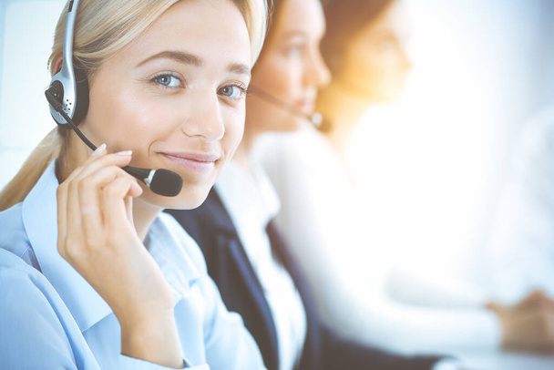 Cheerful smiling business woman with headphones consulting clients. Group of diverse phone operators at work in sunny office.Call center and business people concept - Fotó, kép