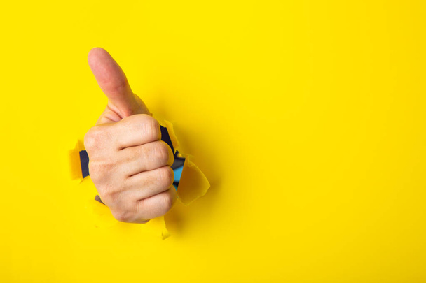 Mano mostrando un signo de pulgar hacia arriba a través de un agujero rasgado en el fondo de papel amarillo. - Foto, imagen