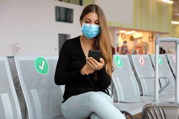 Voyageur femme avec masque chirurgical en utilisant un téléphone intelligent et assis en respectant la distance sociale au salon de l'aéroport - Photo, image