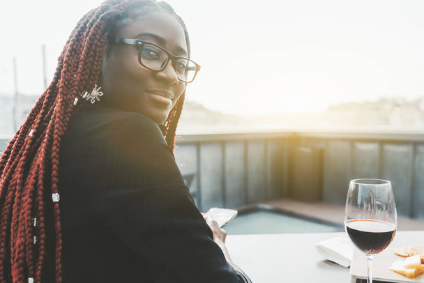 A portrait of a cheerful young black woman in eyeglasses and with braids of maroon color, sitting at a restaurant table outdoors with a glass of wine and phoning; a copy space place on the right - Фото, зображення