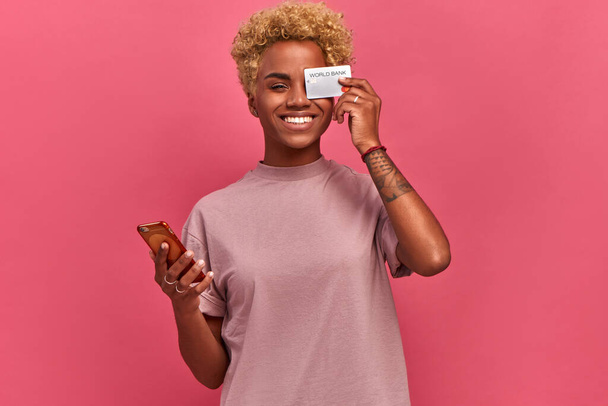 Young dark-skinned woman freelancer holding a bank card and phone on a pink background. - Photo, Image