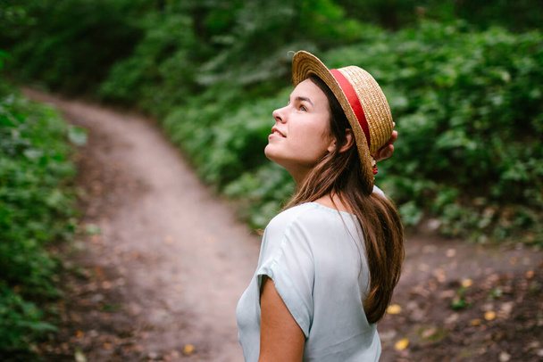 Молодая женщина в соломенной шляпе в летнем лесу. Милая молодая леди, гуляющая по лесу. Красивая белая дама в винтажной шляпе. Студент гуляет на природе в выходной. Девушка ходит в поход после занятий. - Фото, изображение