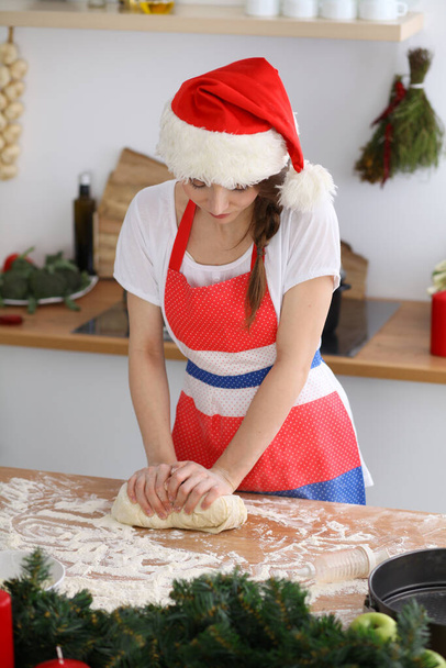 Young brunette woman cooking pizza or handmade pasta while wearing Santa Claus cap in the kitchen. Housewife preparing dough on wooden table - Фото, зображення