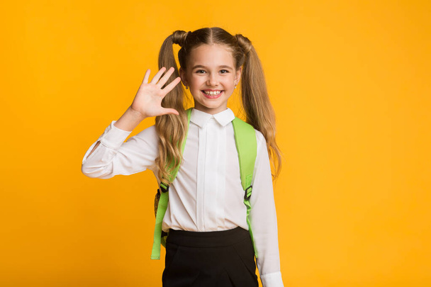 Happy Schoolgirl Waving Hand Smiling To Camera On Yellow Background - Photo, Image