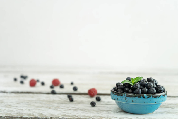 A beautiful blue bowl full of ripe blueberries stands on a light wooden table against a white wall. The bowl is decorated with fresh mint leaves. There are raspberries and blueberries on the table. - 写真・画像