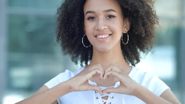 Piękna młoda Afroamerykańska nastolatka uśmiecha się zbyt mocno, składa ręce w znak serca przed nią. Przyjazny etniczny kobieta patrzy w miłość, pojęcie jak, zaufanie, przyjaźń, miłosierdzie, nadzieja.  - Materiał filmowy, wideo