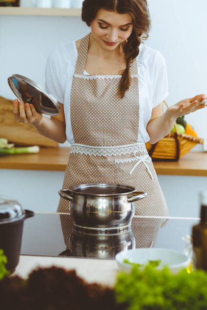 Young brunette woman cooking soup in kitchen. Housewife holding wooden spoon in her hand. Food and health concept - Фото, изображение