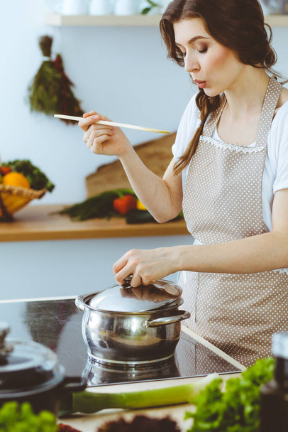 Молодая брюнетка готовит суп на кухне. Домохозяйка держит в руке деревянную ложку. Концепция питания и здоровья - Фото, изображение