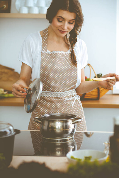 Young brunette woman cooking soup in kitchen. Housewife holding wooden spoon in her hand. Food and health concept - Photo, image